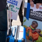 A laid off USAID employee holds a poster of her work, oath of office, and a suitcase of personal belongings after being given 15 minutes to clear out of the USAID headquarters in Washington DC.