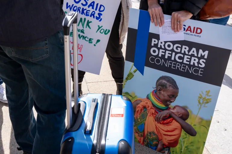 A laid off USAID employee holds a poster of her work, oath of office, and a suitcase of personal belongings after being given 15 minutes to clear out of the USAID headquarters in Washington DC.